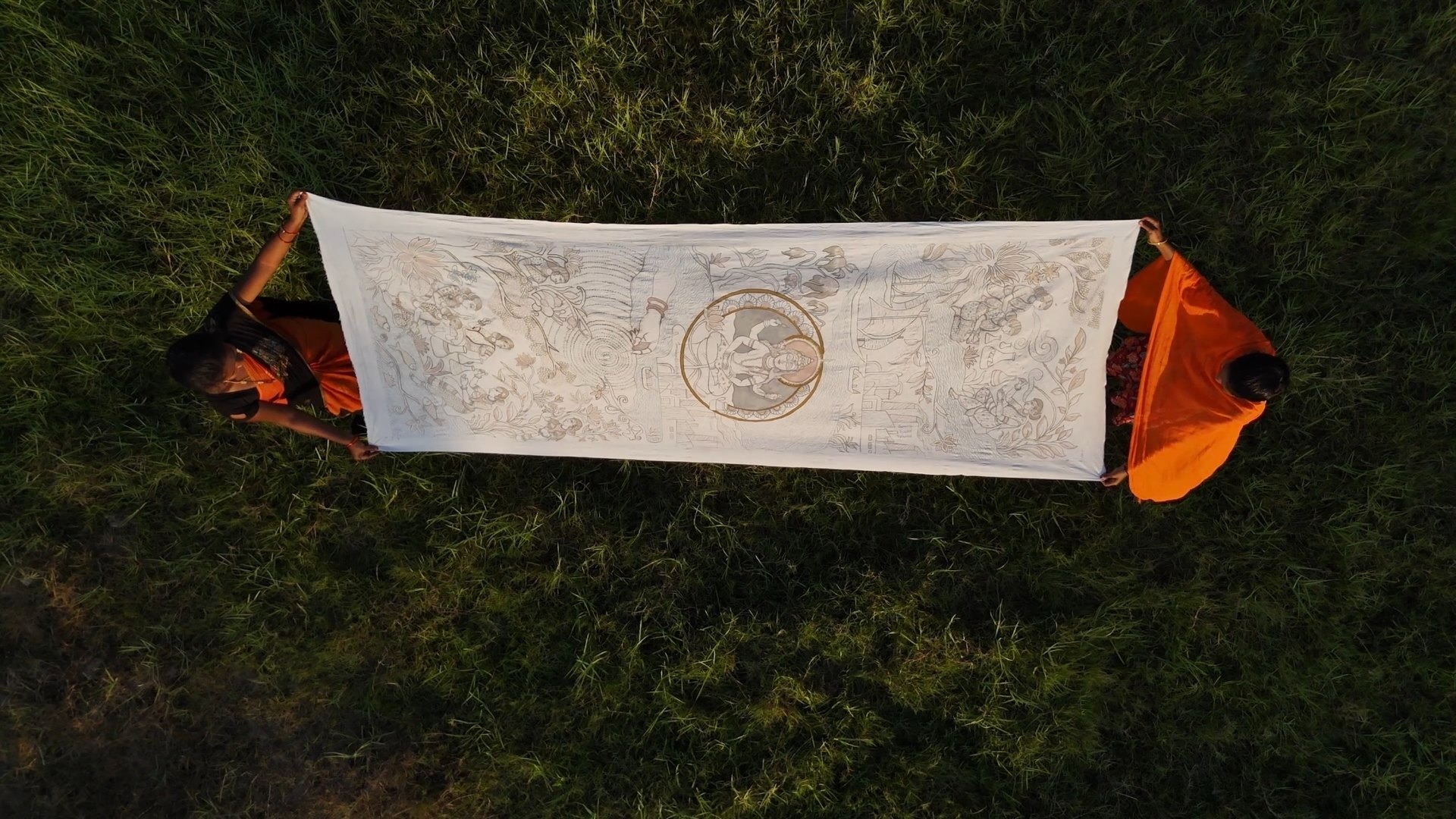 Two artisans holding a long painted kalamkari cloth open over grass, seen from above
