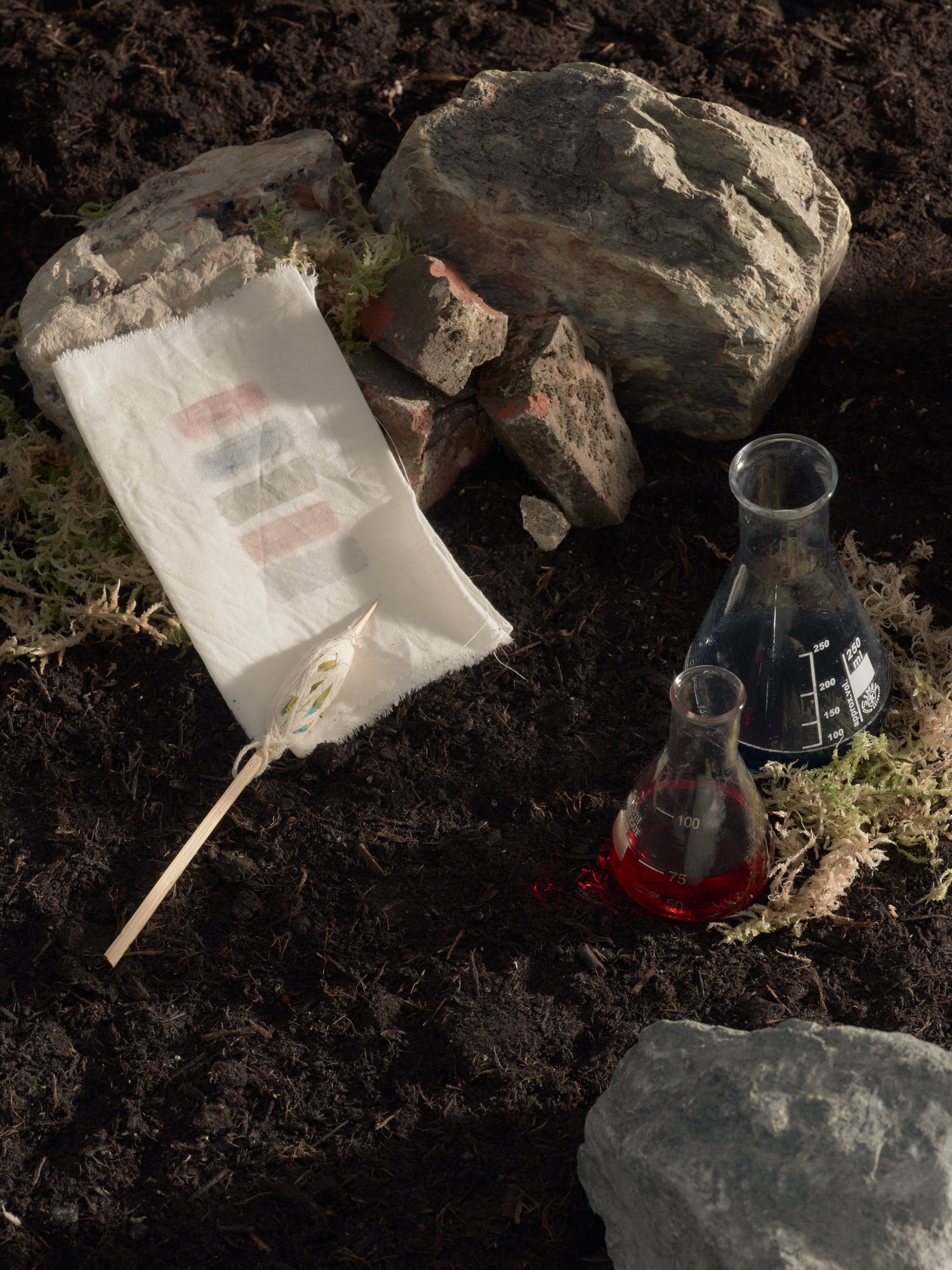 Flasks of red and dark pigment set in soil beside dyed cloth and a kalam